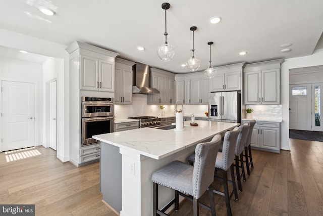 kitchen with a kitchen island with sink, wall chimney range hood, light hardwood / wood-style flooring, sink, and appliances with stainless steel finishes