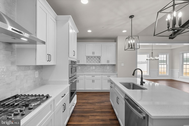 kitchen with wall chimney range hood, an island with sink, sink, white cabinetry, and appliances with stainless steel finishes