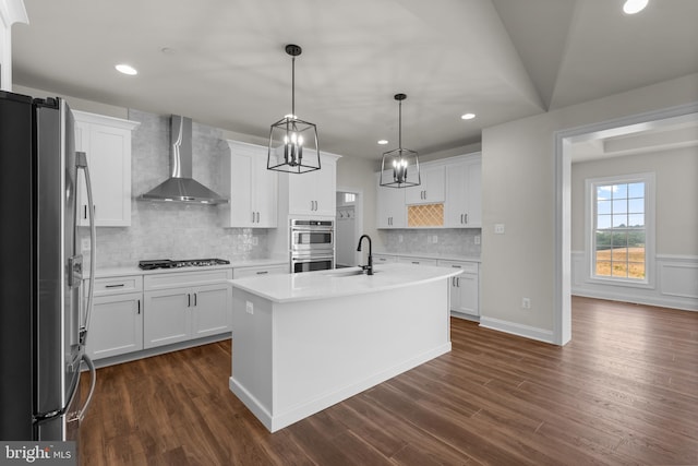 kitchen featuring wall chimney range hood, an island with sink, white cabinets, pendant lighting, and appliances with stainless steel finishes