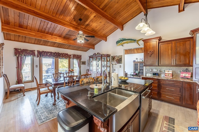 kitchen with light wood-type flooring, an island with sink, ceiling fan, dark stone counters, and stainless steel dishwasher