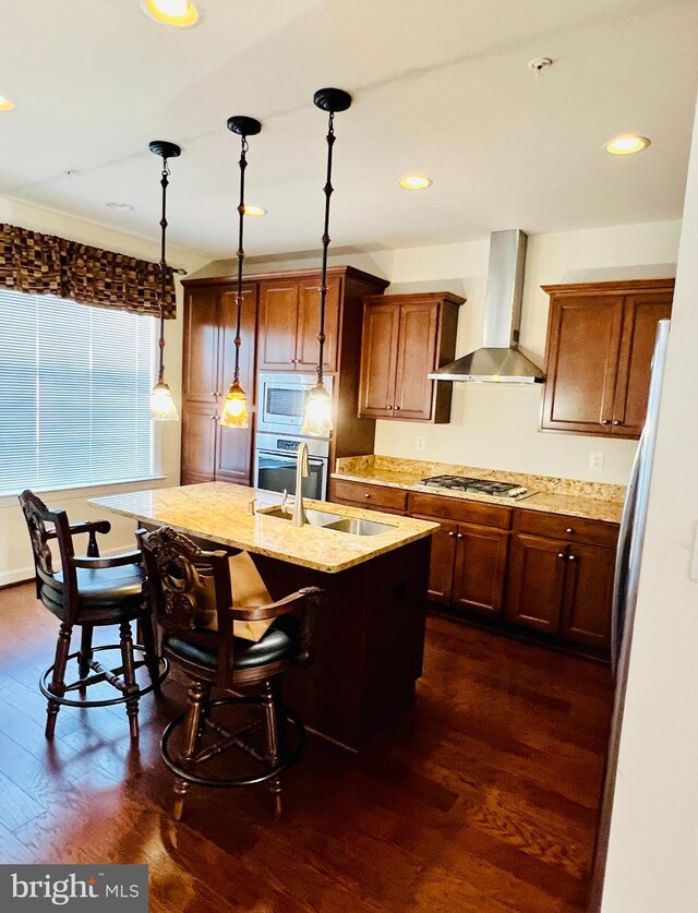 kitchen with wall chimney exhaust hood, stainless steel appliances, dark hardwood / wood-style floors, an island with sink, and decorative light fixtures