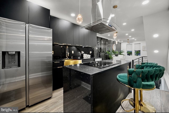 kitchen with stainless steel fridge, island exhaust hood, backsplash, black electric stovetop, and decorative light fixtures