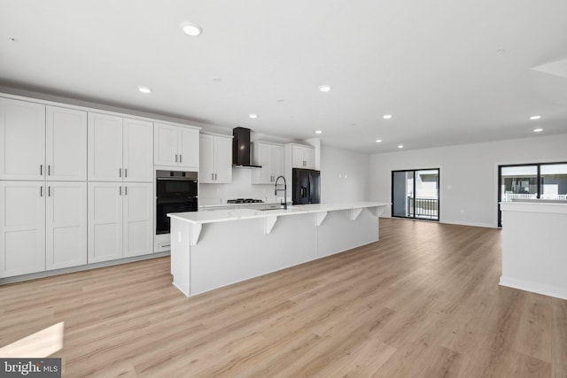 kitchen featuring a spacious island, wall chimney range hood, black appliances, a breakfast bar, and light wood-type flooring