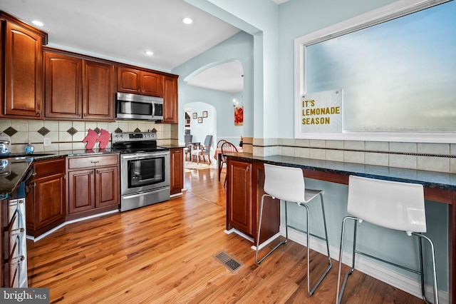 kitchen with decorative backsplash, stainless steel appliances, dark stone counters, and light hardwood / wood-style floors