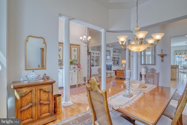 dining area featuring a high ceiling, a fireplace, light wood-style floors, decorative columns, and an inviting chandelier