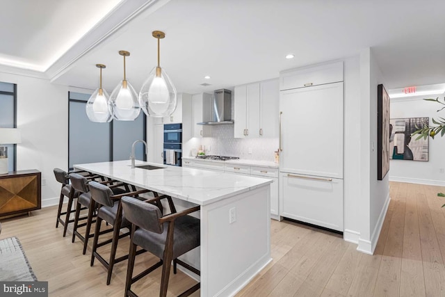 kitchen featuring a kitchen breakfast bar, wall chimney exhaust hood, paneled built in fridge, white cabinets, and a kitchen island with sink