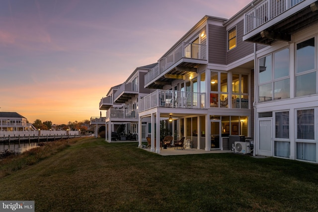 back house at dusk with a balcony, a patio, and a lawn