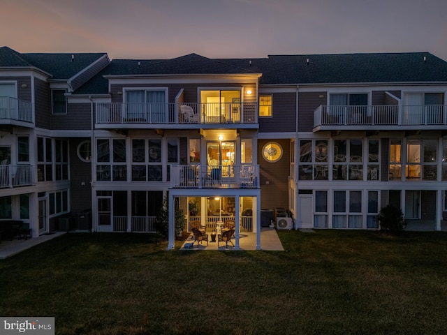 back house at dusk with a balcony, a patio, central AC, and a lawn