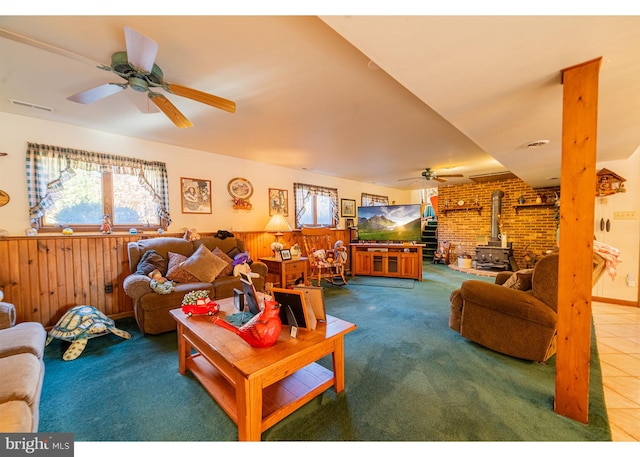 tiled living room featuring a wood stove, wood walls, ceiling fan, and brick wall