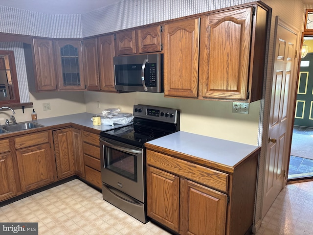 kitchen with a sink, brown cabinetry, stainless steel appliances, light floors, and glass insert cabinets