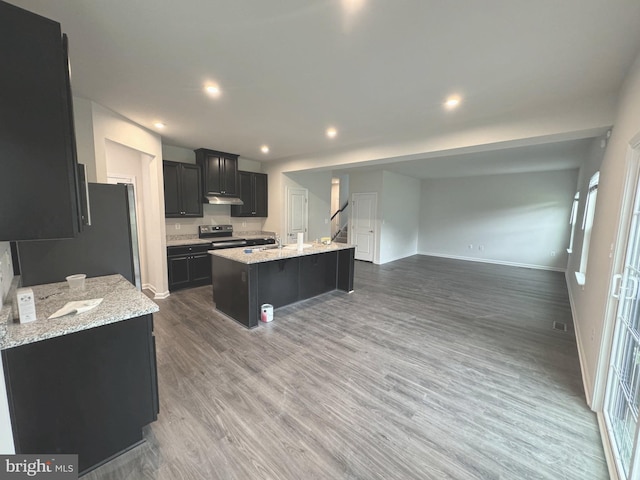 kitchen featuring under cabinet range hood, stainless steel electric stove, wood finished floors, a kitchen island with sink, and baseboards