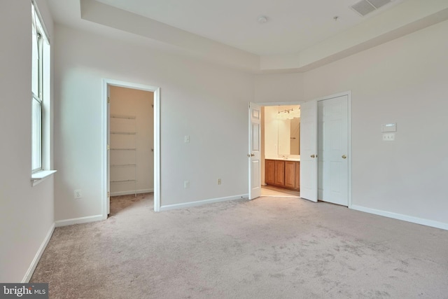 unfurnished bedroom featuring a spacious closet, a raised ceiling, light colored carpet, and ensuite bath