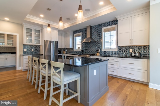 kitchen featuring wall chimney exhaust hood, a tray ceiling, light hardwood / wood-style floors, and a center island with sink