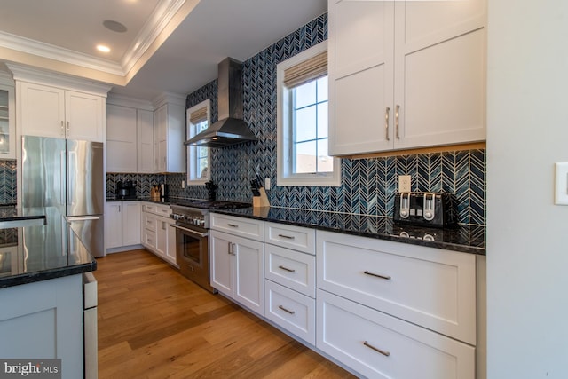 kitchen with wall chimney range hood, appliances with stainless steel finishes, light wood-type flooring, white cabinets, and dark stone countertops