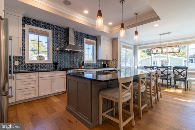 kitchen with wall chimney range hood, an island with sink, white cabinetry, and light hardwood / wood-style floors