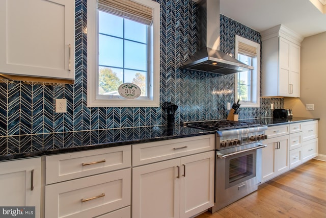 kitchen with white cabinetry, wall chimney exhaust hood, high end stove, dark stone countertops, and light hardwood / wood-style flooring