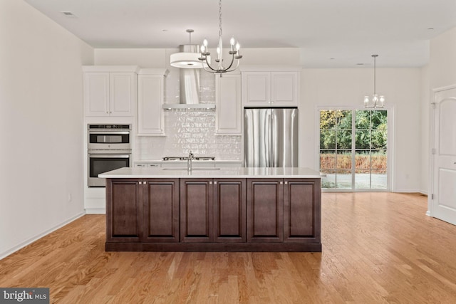kitchen with light countertops, an island with sink, stainless steel appliances, wall chimney range hood, and a chandelier