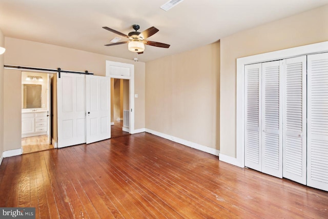 unfurnished bedroom featuring ensuite bath, ceiling fan, wood-type flooring, a barn door, and a closet