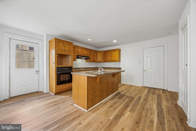 kitchen with light hardwood / wood-style floors, light stone counters, a kitchen bar, oven, and a kitchen island with sink