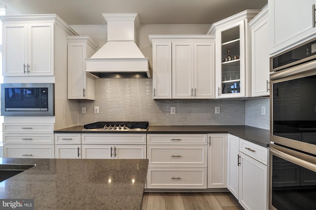 kitchen featuring light hardwood / wood-style floors, dark stone counters, white cabinets, custom range hood, and appliances with stainless steel finishes