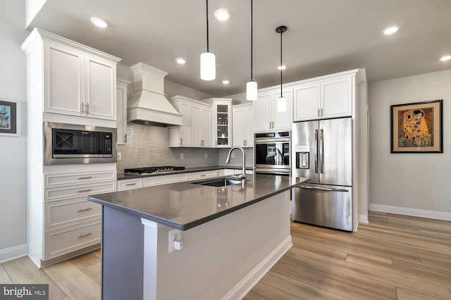 kitchen with stainless steel appliances, sink, custom range hood, decorative light fixtures, and an island with sink