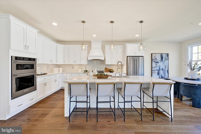 kitchen with custom exhaust hood, white cabinetry, stainless steel appliances, and an island with sink