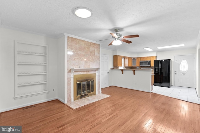 unfurnished living room featuring ceiling fan, built in features, a textured ceiling, a tiled fireplace, and light wood-type flooring