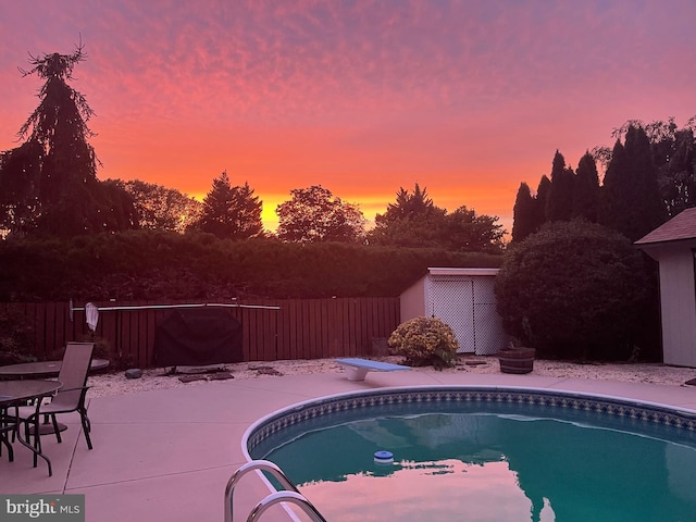 pool at dusk featuring a diving board, a patio, and a shed