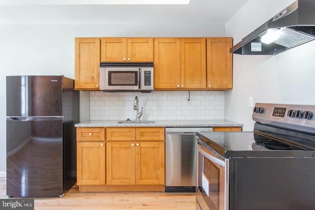 kitchen with ventilation hood, light stone counters, light wood-type flooring, appliances with stainless steel finishes, and sink