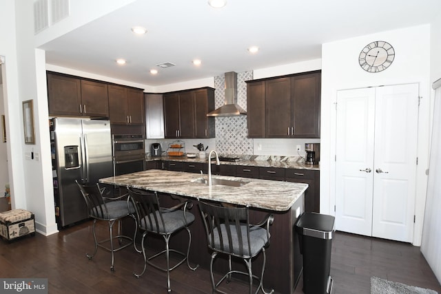 kitchen with stainless steel appliances, wall chimney range hood, dark hardwood / wood-style flooring, an island with sink, and a breakfast bar