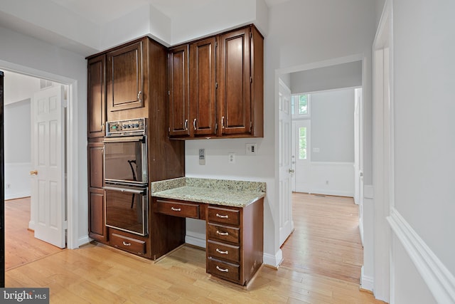 kitchen featuring multiple ovens, dark brown cabinets, light stone counters, built in desk, and light wood-type flooring