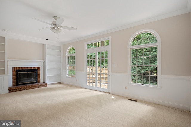 unfurnished living room with crown molding, ceiling fan, a fireplace, built in shelves, and light colored carpet