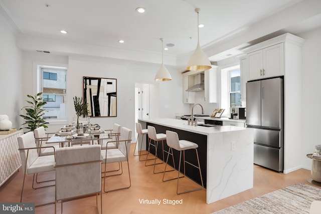 kitchen featuring wall chimney range hood, a breakfast bar, pendant lighting, white cabinetry, and stainless steel fridge