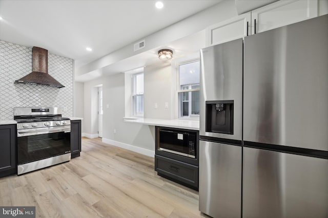 kitchen with tasteful backsplash, white cabinetry, appliances with stainless steel finishes, wall chimney exhaust hood, and light hardwood / wood-style flooring