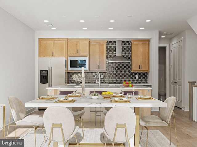 kitchen with white microwave, a center island with sink, wall chimney range hood, stainless steel refrigerator with ice dispenser, and decorative backsplash