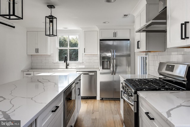 kitchen with wall chimney range hood, appliances with stainless steel finishes, hanging light fixtures, sink, and light hardwood / wood-style floors