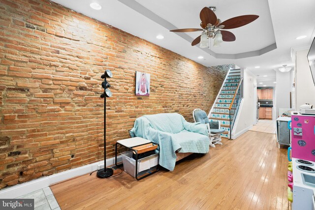 living area featuring light hardwood / wood-style flooring, a raised ceiling, ceiling fan, and brick wall