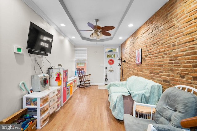 rec room featuring ceiling fan, brick wall, a tray ceiling, and light hardwood / wood-style flooring