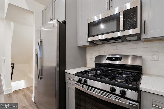 kitchen featuring light wood-type flooring, appliances with stainless steel finishes, gray cabinets, light stone countertops, and decorative backsplash