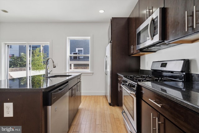 kitchen featuring stainless steel appliances, light hardwood / wood-style floors, sink, dark stone counters, and an island with sink