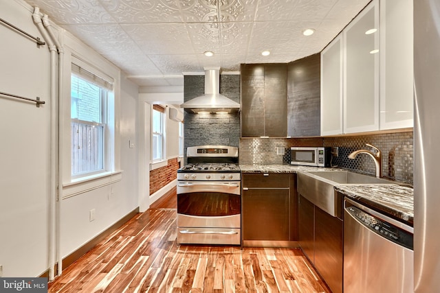 kitchen featuring stainless steel appliances, wall chimney range hood, light stone countertops, white cabinetry, and light hardwood / wood-style flooring