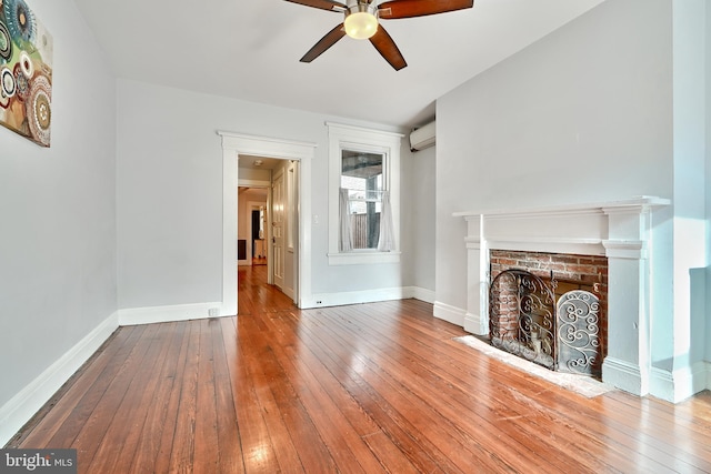 unfurnished living room featuring a wall mounted AC, hardwood / wood-style floors, a brick fireplace, and ceiling fan