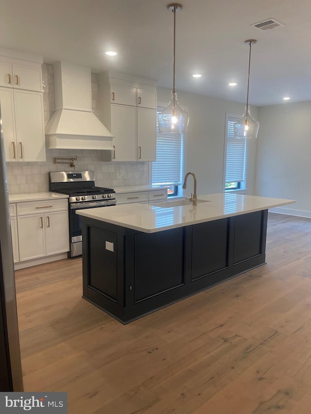 kitchen with premium range hood, an island with sink, white cabinets, decorative light fixtures, and stainless steel gas stove