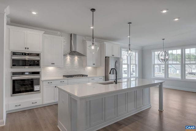 kitchen with a center island with sink, wall chimney exhaust hood, hanging light fixtures, sink, and appliances with stainless steel finishes