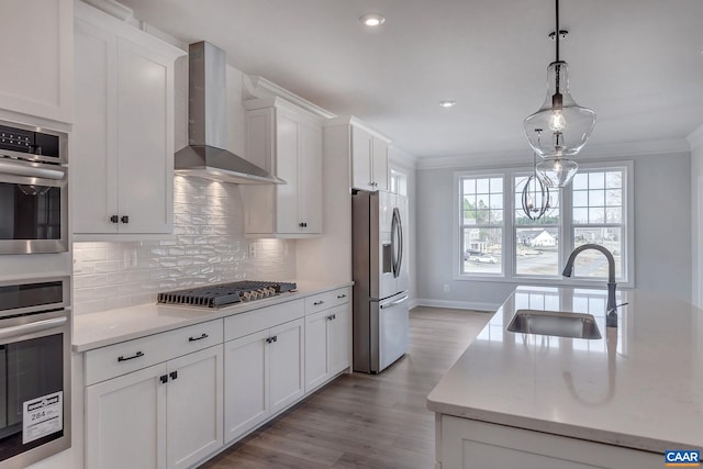 kitchen with sink, appliances with stainless steel finishes, hanging light fixtures, white cabinets, and wall chimney range hood