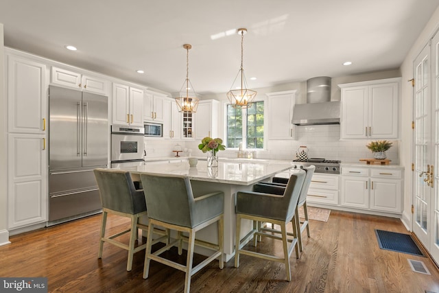 kitchen with wall chimney exhaust hood, wood-type flooring, built in appliances, white cabinets, and a center island