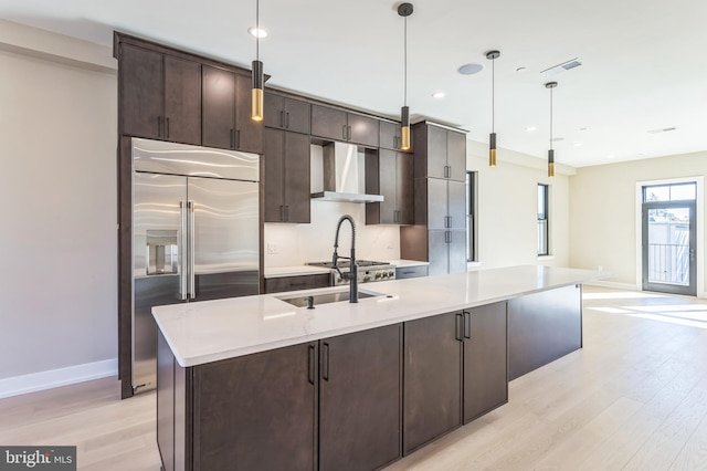 kitchen with appliances with stainless steel finishes, a center island with sink, hanging light fixtures, and wall chimney exhaust hood