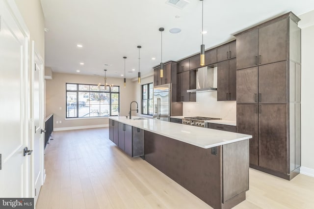 kitchen featuring wall chimney exhaust hood, hanging light fixtures, light hardwood / wood-style flooring, a center island with sink, and appliances with stainless steel finishes