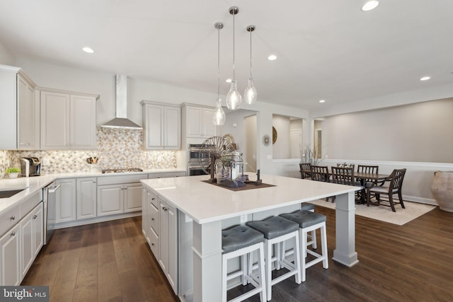 kitchen with wall chimney range hood, white cabinetry, stainless steel appliances, a kitchen island, and decorative light fixtures