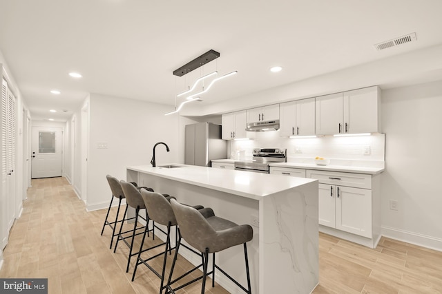 kitchen featuring stainless steel appliances, light hardwood / wood-style floors, white cabinets, hanging light fixtures, and an island with sink